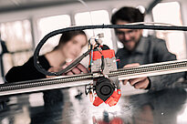 A machine is in focus in the foreground, two students, out of focus, are observing it in the background